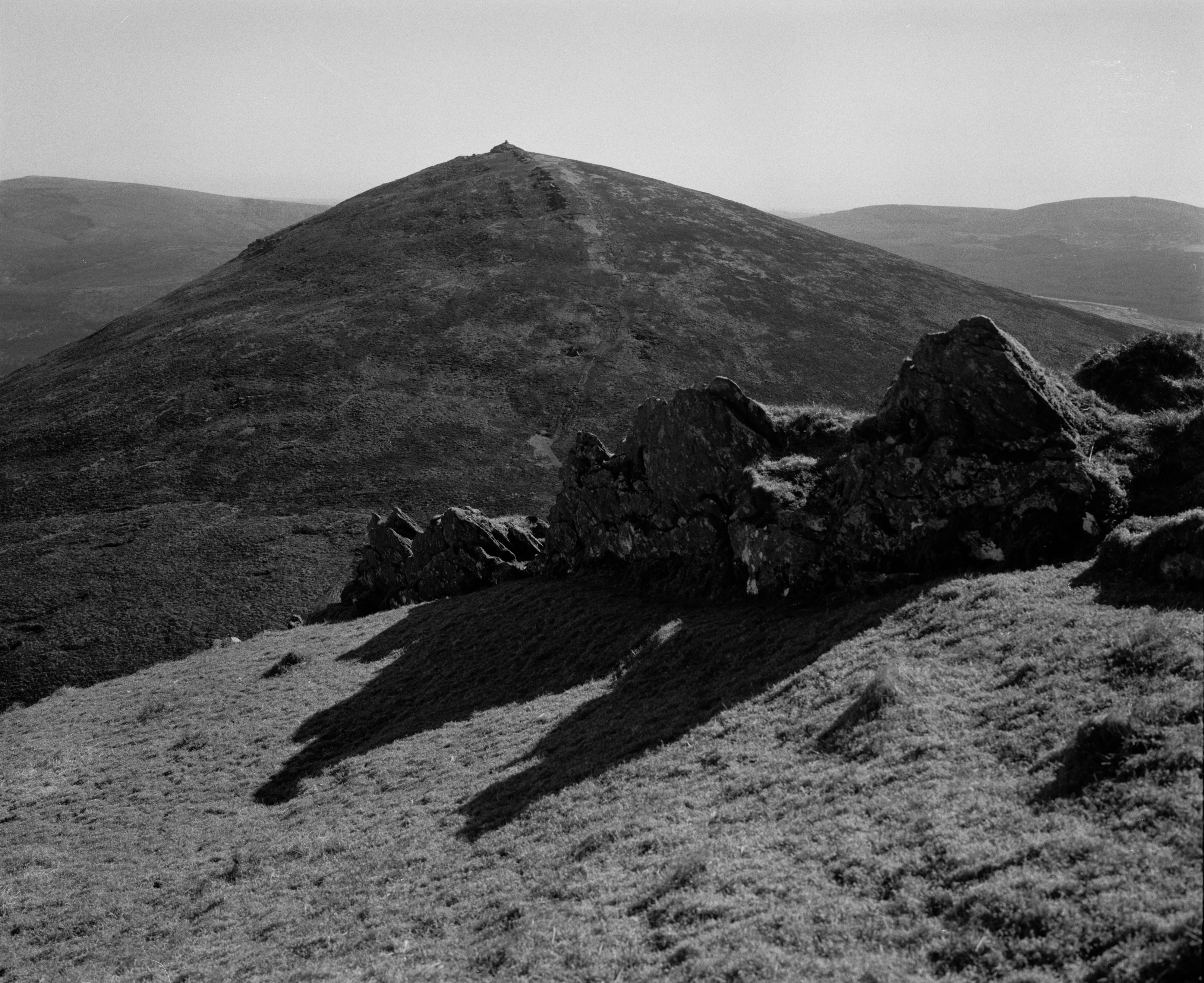 A black and white photograph of a large, rounded mountain under clear skies. A faint path ascends the slope toward a rocky summit. In the foreground, jagged rocks cast long shadows over the grassy terrain, adding contrast and depth to the scene. Rolling hills stretch out in the background
