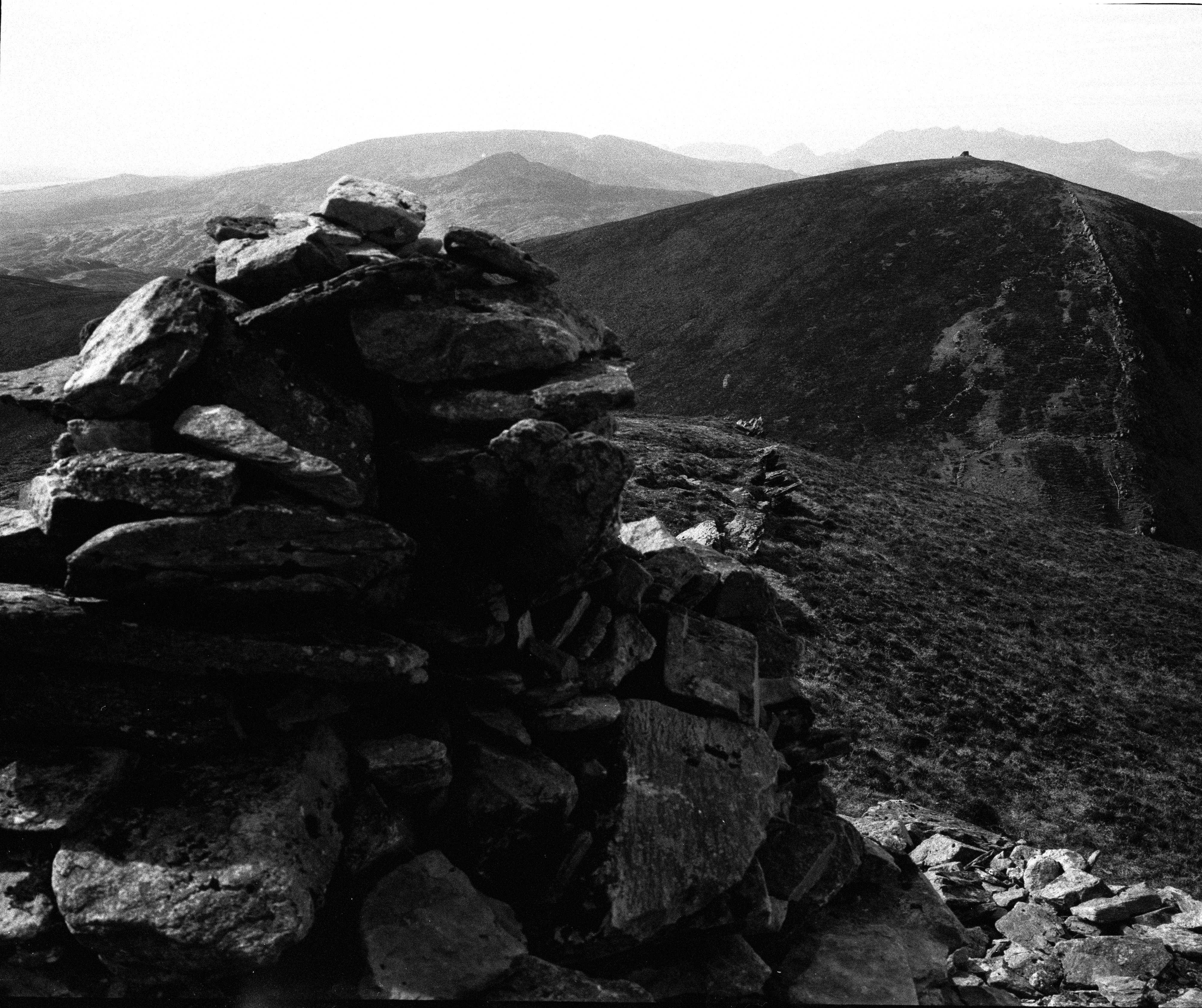 A black and white photograph of a mountainous landscape, featuring a cairn made of stacked stones in the foreground. Rolling hills and rugged peaks stretch into the background, with a steep path visible along the ridge of a prominent hill. The atmosphere is stark and dramatic, emphasizing the natural textures and contours of the terrain.