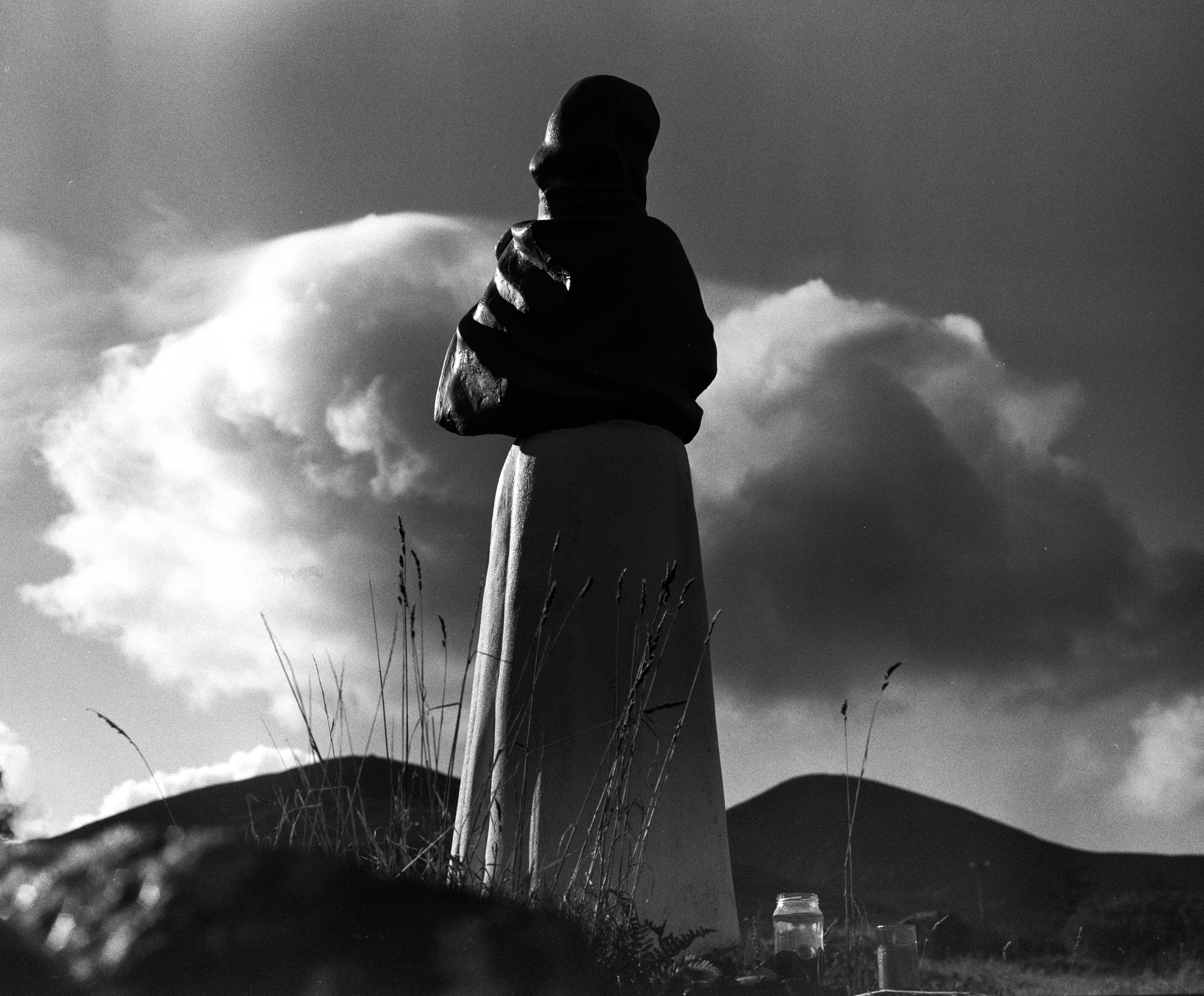 A black and white photograph of a statue of a robed figure, viewed from behind and silhouetted against a dramatic sky with large, billowing clouds. The figure stands in a grassy field with the Pap mountains in the background. In the foreground, two small jars, containing candles or offerings, rest on the ground near tall blades of grass
