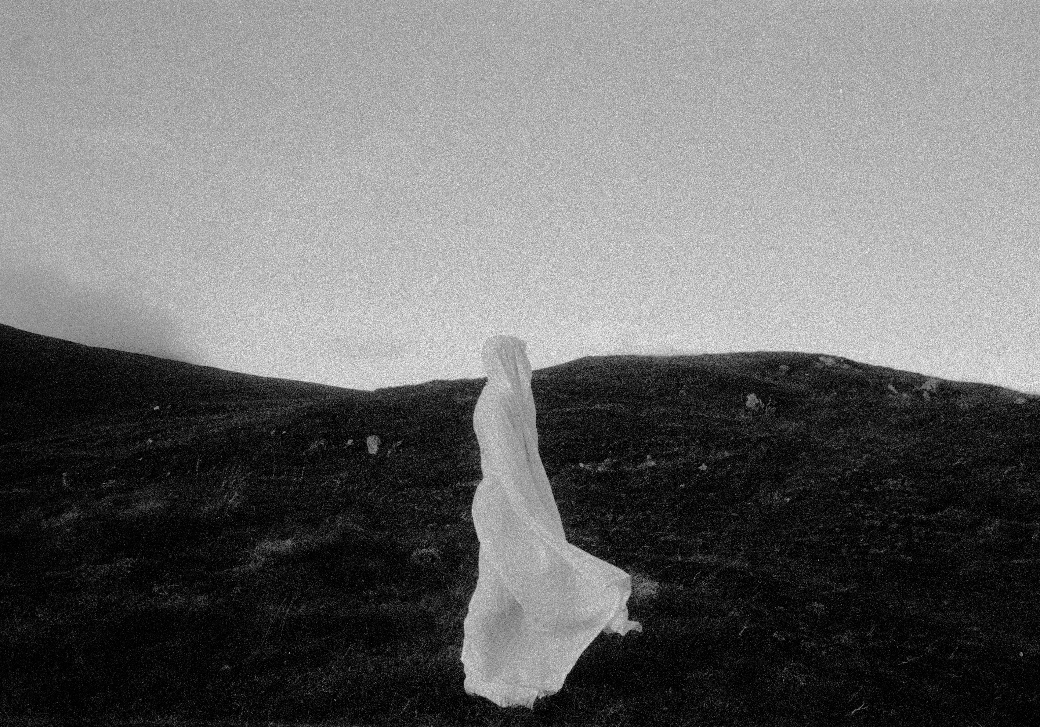 A black-and-white photograph of a solitary figure draped in a flowing white cloth standing on a barren hillside. The figure faces to the right, blending into the stark, windswept landscape of rolling hills and dark terrain under a vast, empty sky. The ethereal appearance creates a haunting, ghostlike atmosphere.