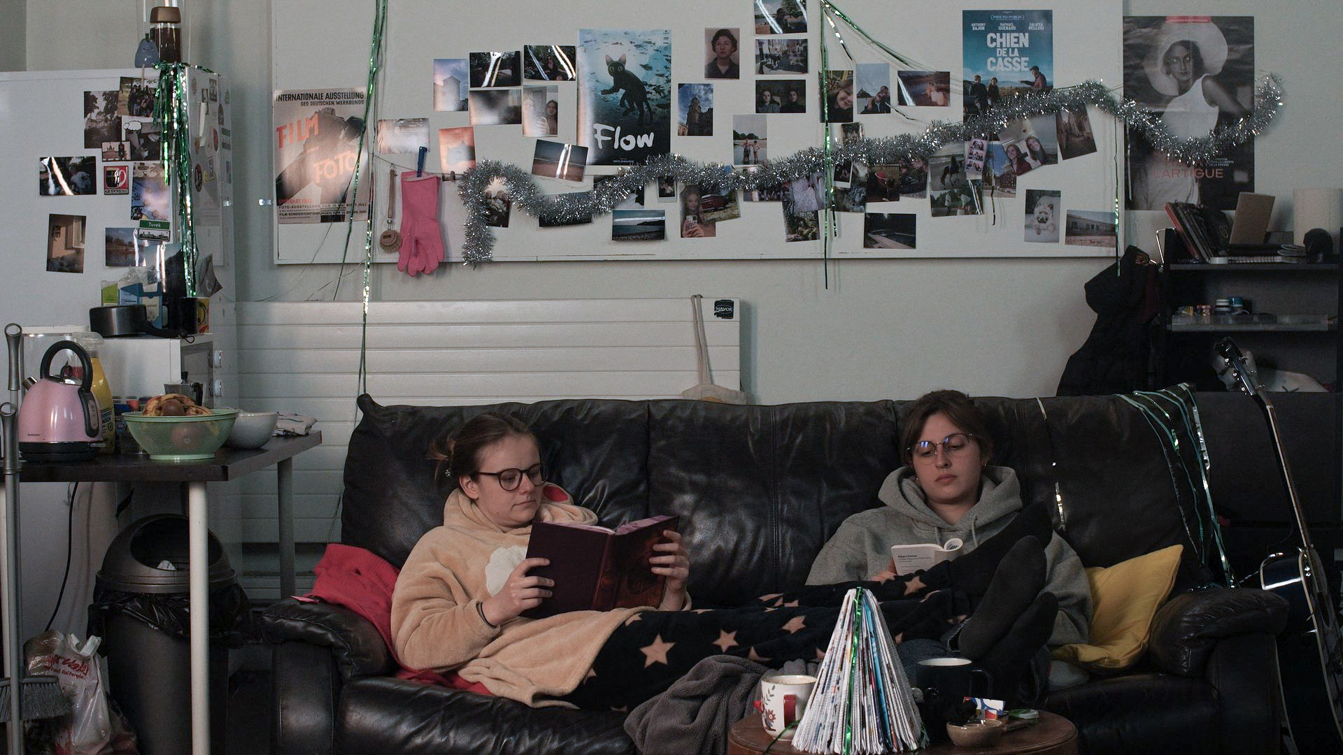 The photograph shows two young women sitting on a black leather sofa. They are sitting in the middle of a messy living room. The young woman on the left is reading a book. She is wearing reindeer pyjamas and glasses. The woman on the right, wearing a grey hoodie and glasses, is also reading a book. They are not looking at each other, but sharing a moment together.