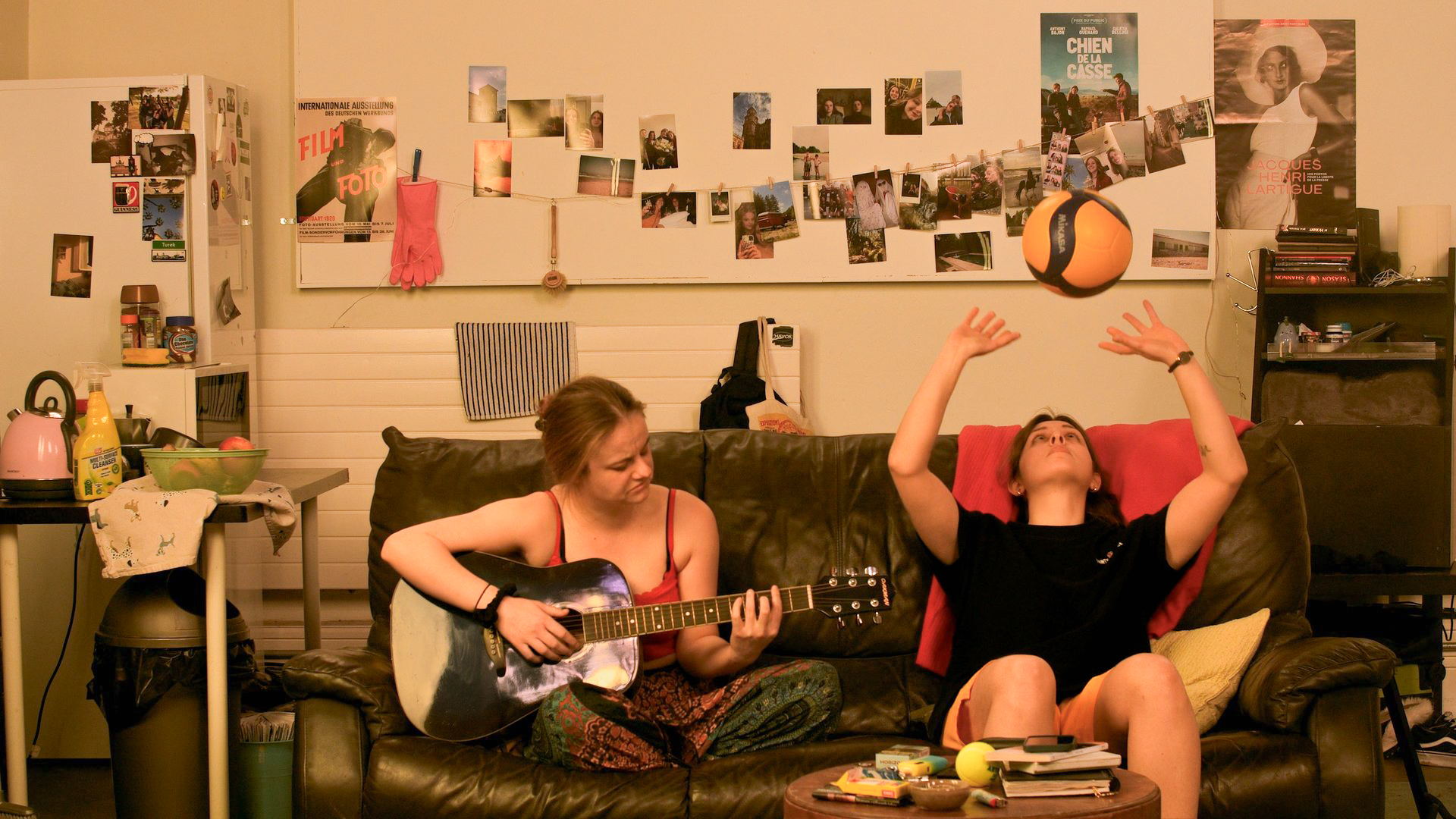 The photograph shows two young women sitting on a black leather sofa. They are sitting in the middle of a messy living room. The young woman on the left is playing her guitar. She is wearing a red t-shirt with straps and sirwal pants. The woman on the right, wearing an oversized black T-shirt and yellow shorts, is bouncing a volleyball in her hands. They don't look at each other, but share a moment together.  