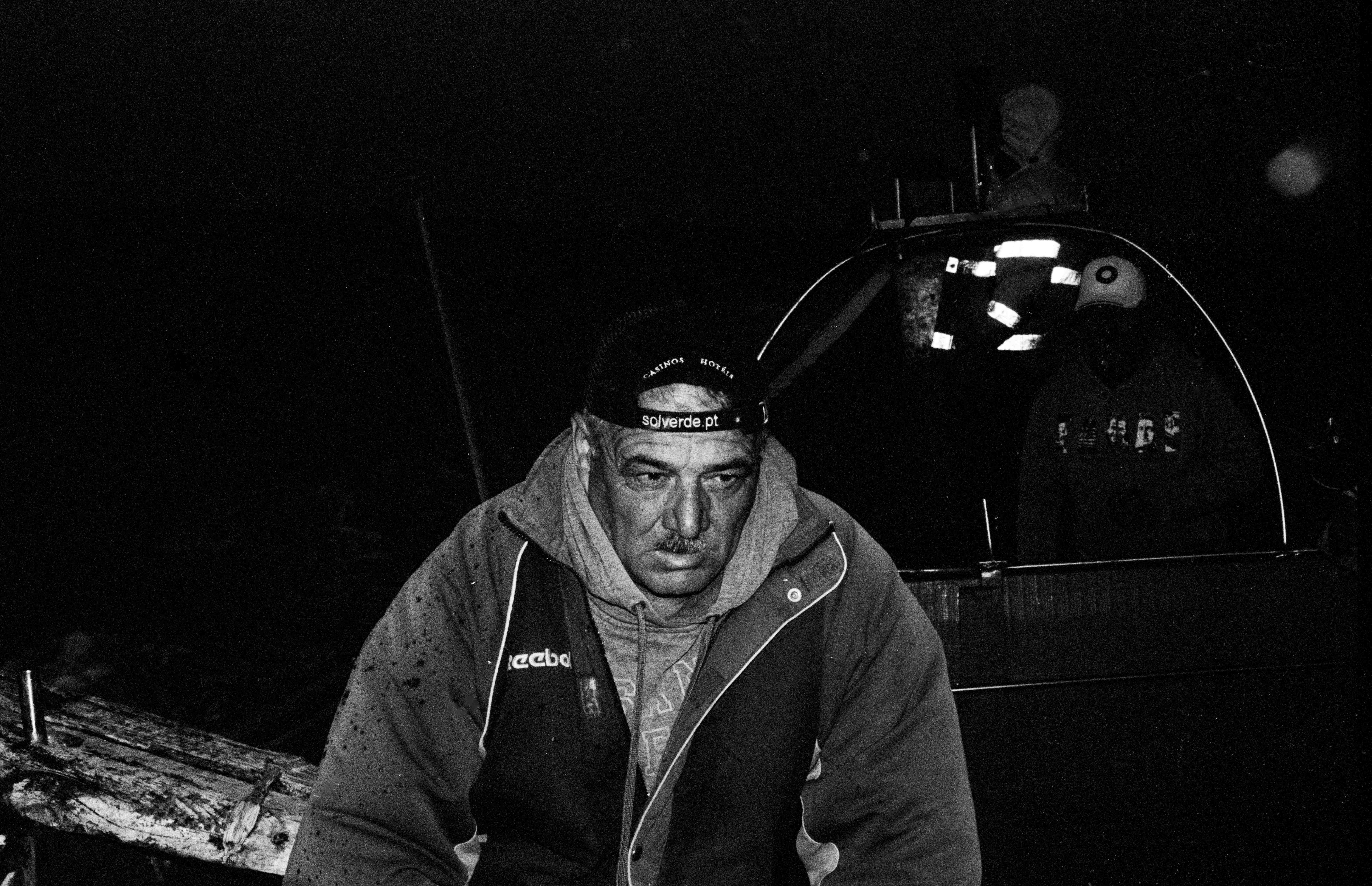 Portrait of a fisherman called Zé, the Image was shot in black and white and was taken at 6AM on the Boat, the background is dark and the rest of the Fishermen are barely visible.