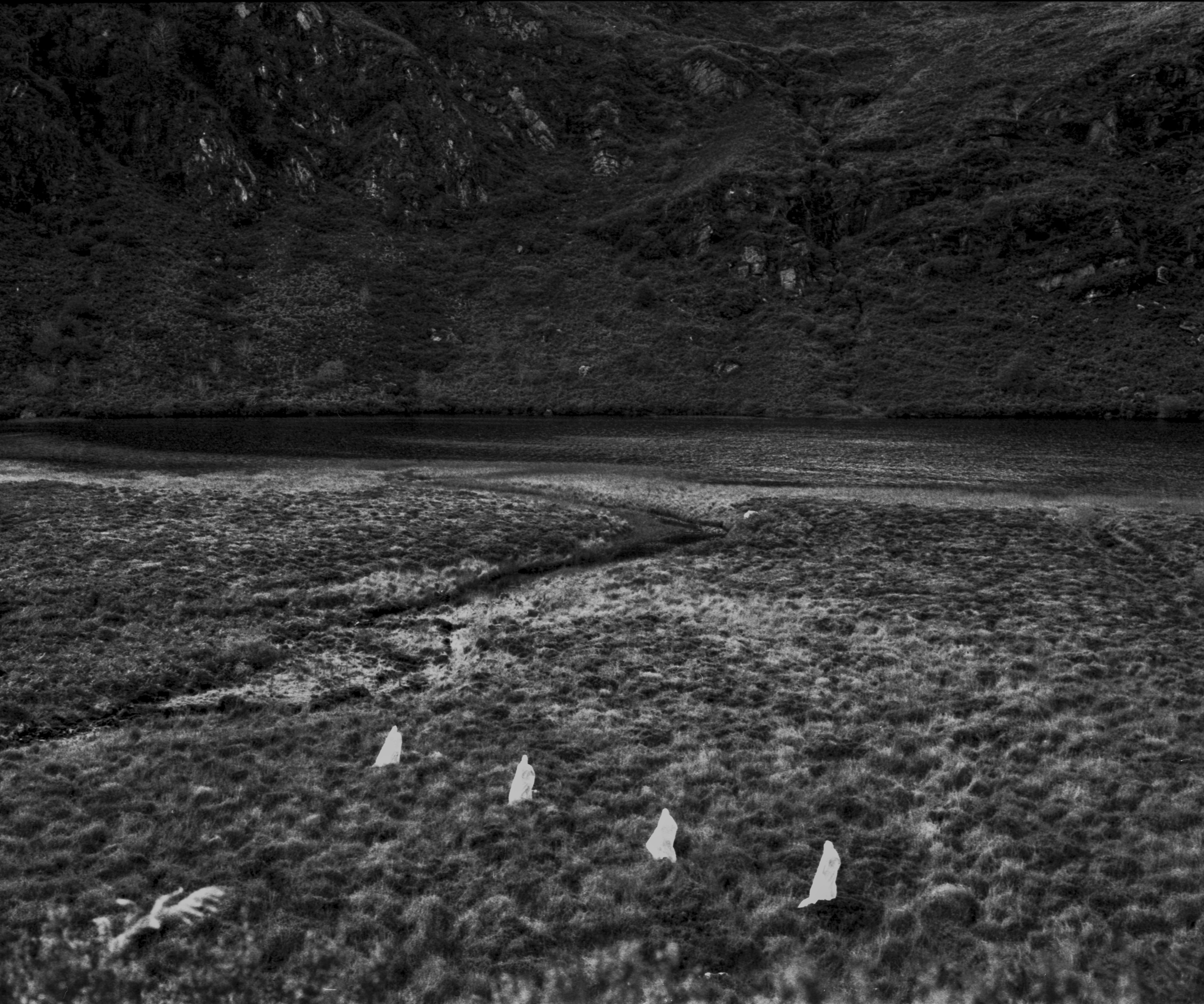 A black and white photograph of a grassy moorland landscape with a narrow stream winding through the terrain toward a dark, still body of water in the background. The far side of the water is bordered by a steep, rocky hillside covered in dense vegetation. In the foreground, four small white figures are staked into the ground in a line, contrasting with the dark grass.
