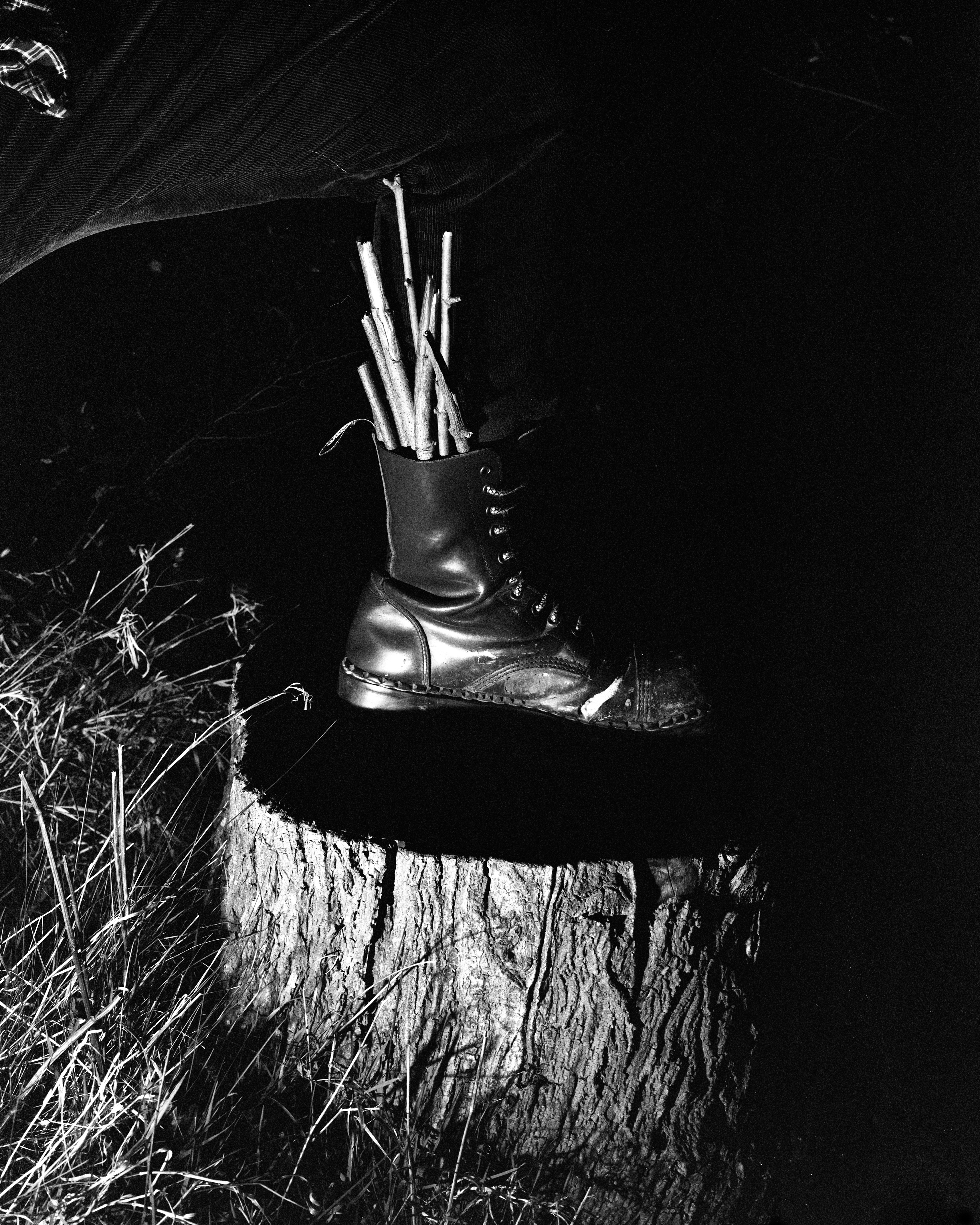 A black and white photo showing a person’s leg wearing a shiny combat boot, with several small sticks protruding from inside the boot. The boot is resting on top of a tree stump at night, surrounded by tall grass. The scene is dramatically lit, creating strong contrasts and shadows.