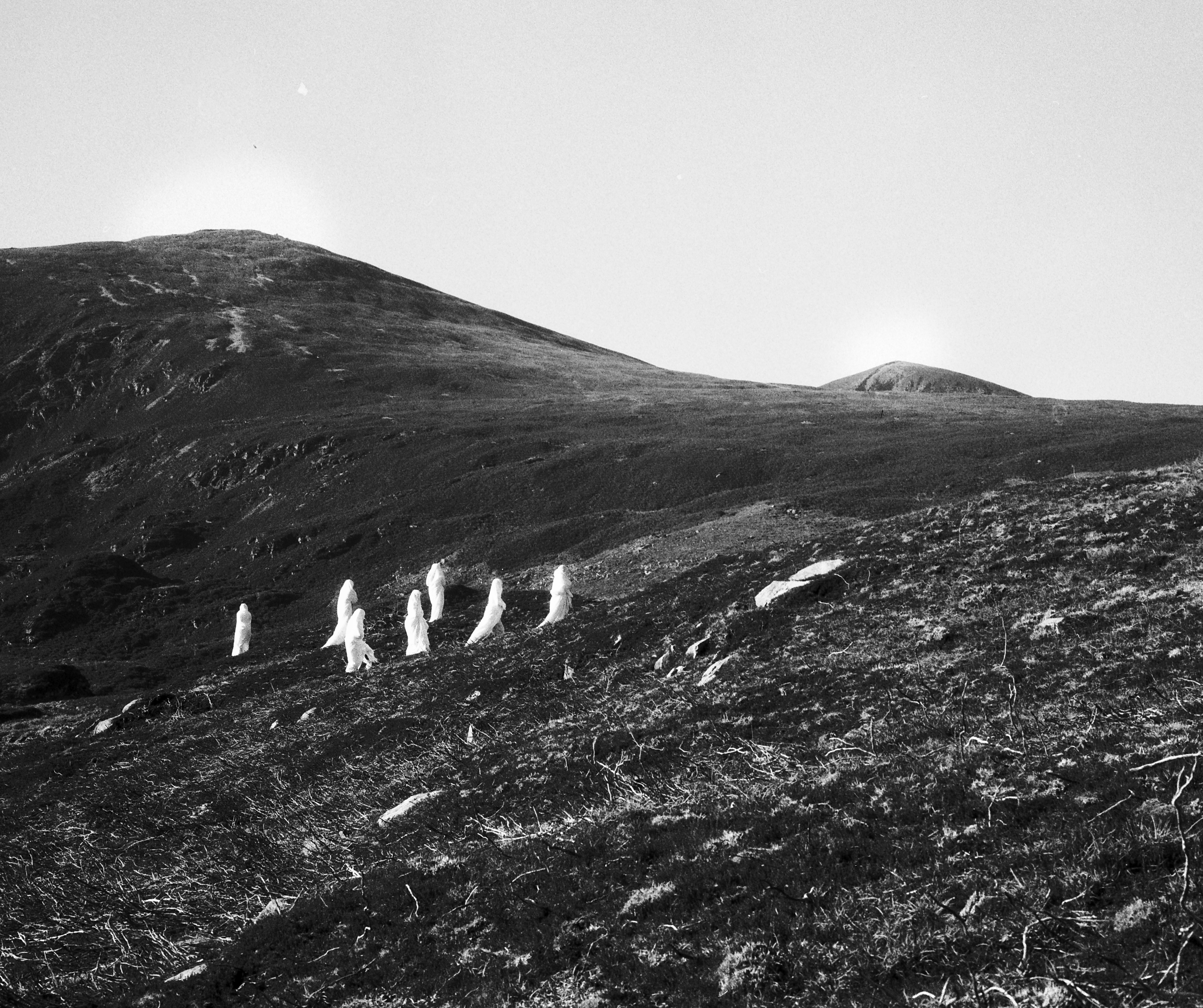 Black and white photograph of a barren, rocky hillside with a group of figures dressed in white, sheet-like garments walking in a loose formation across the slope. In the background, two rounded hills (Paps) rise under a clear sky, with soft halos of light near their peaks. The landscape is stark and desolate, evoking a mysterious or surreal atmosphere