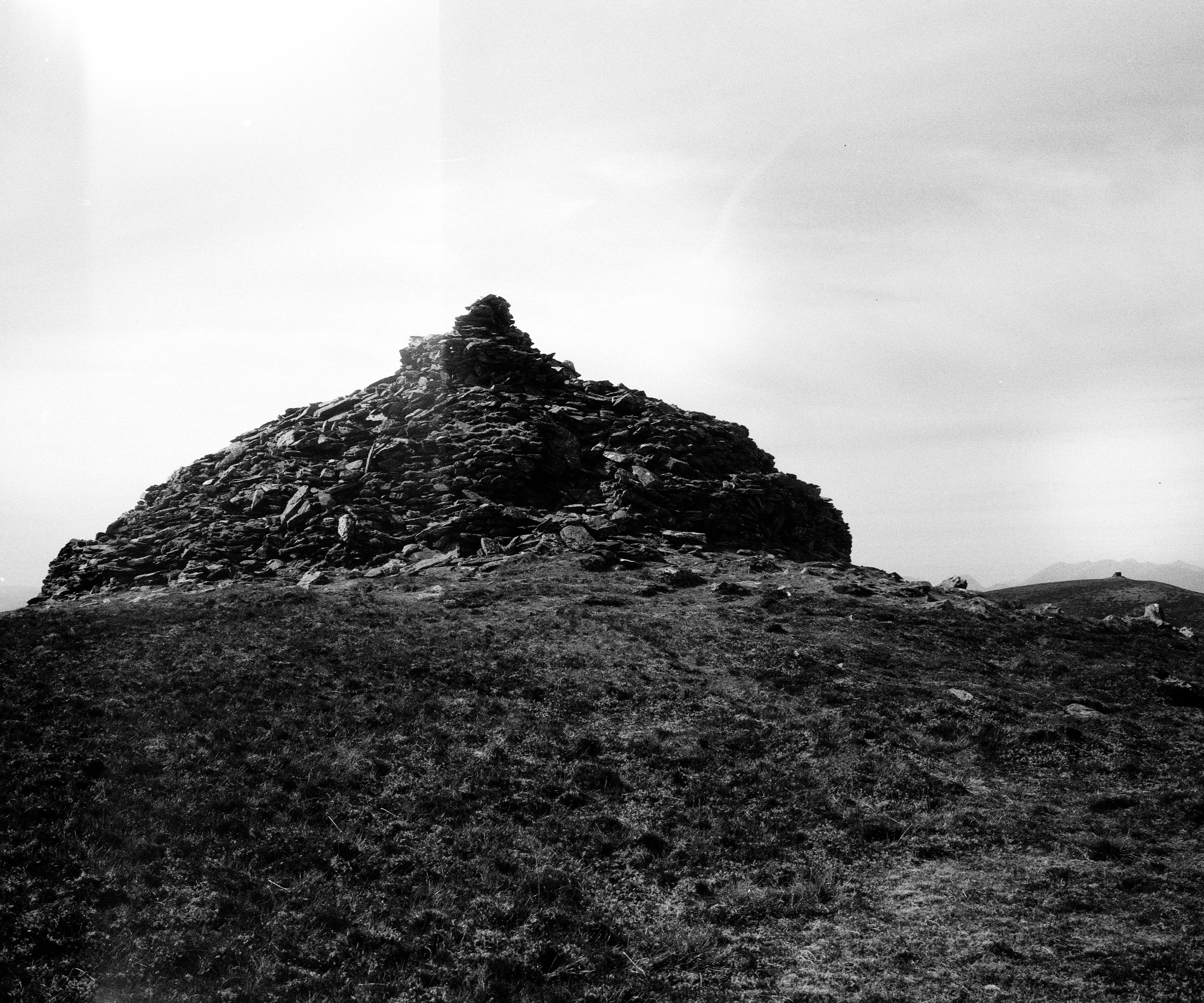 Black and white photograph of a large cairn made of stacked stones, situated on a grassy mountaintop. The cairn rises in a conical shape, its dark texture contrasting against the bright, overcast sky. Distant  parallel hill of mountain peak with similar structure is visible on the horizon, contributing to the importance of the location  with a clear feeling isolation and elevation.
