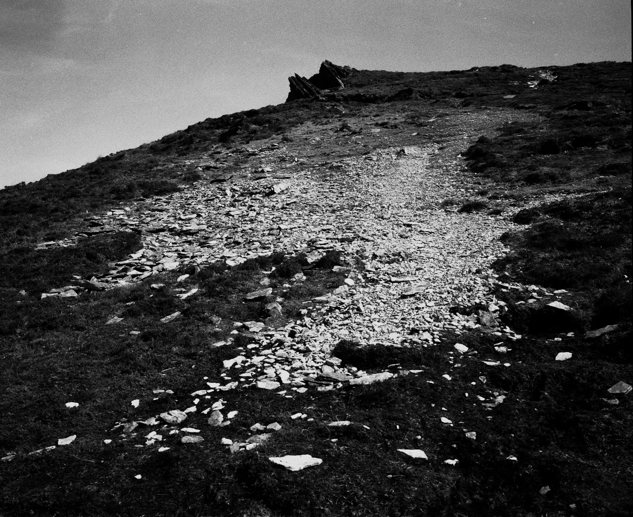 A black-and-white photograph of a rugged hillside with a stony, uneven path made of loose rocks leading upward. The terrain is sparsely covered with short grass and patches of dark earth. Jagged rock formations jut out near the hilltop in the distance, under a lightly clouded sky, adding a dramatic focal point to the stark landscape