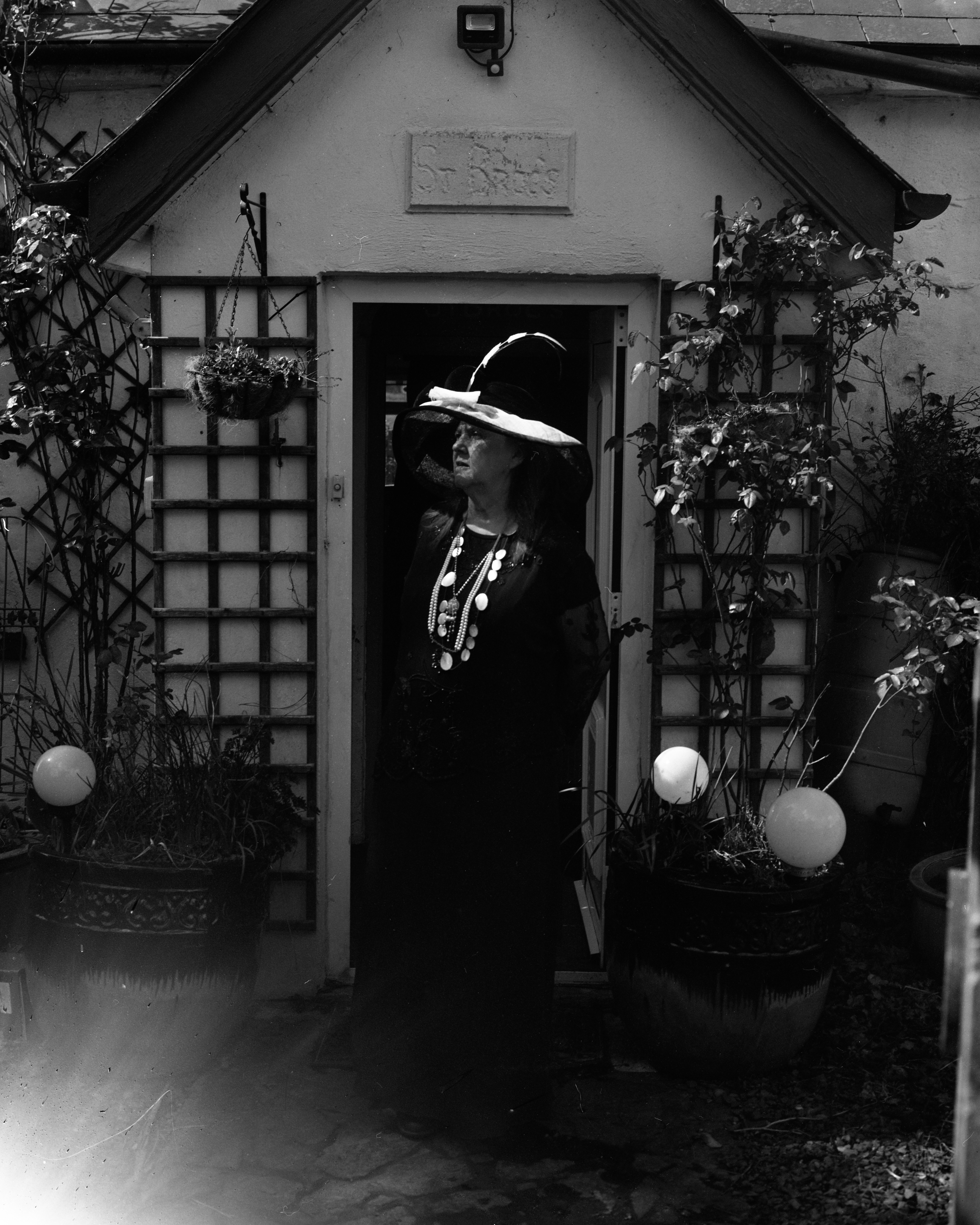 A black and white photograph of a person standing in front of a small house entrance with a peaked roof. The person is dressed in a long, dark gown with a large necklace and a wide-brimmed hat adorned with a feather. The doorway is framed by trellises with climbing plants and large potted plants on either side, each with a round decorative ornament. A light leak creates a bright flare in the lower left corner of the image, adding a dramatic effect. The sign above the door reads "St Brocs"