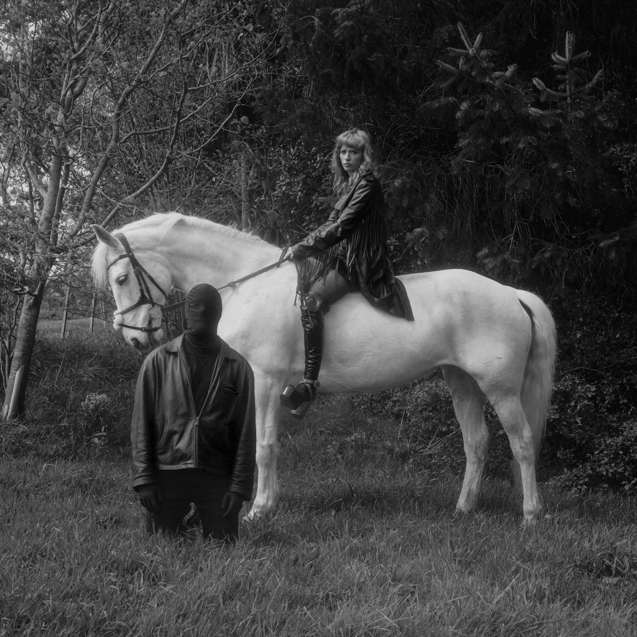 This black and white medium format photograph depicts a large white horse standing perpendicular to the camera. A femdom dressed in leather sits bareback on the horse, looking at the viewer. A masked submissive man in all black and leather kneels on the grass by the horses shoulder, facing the camera. They are surrounded by trees and foliage.