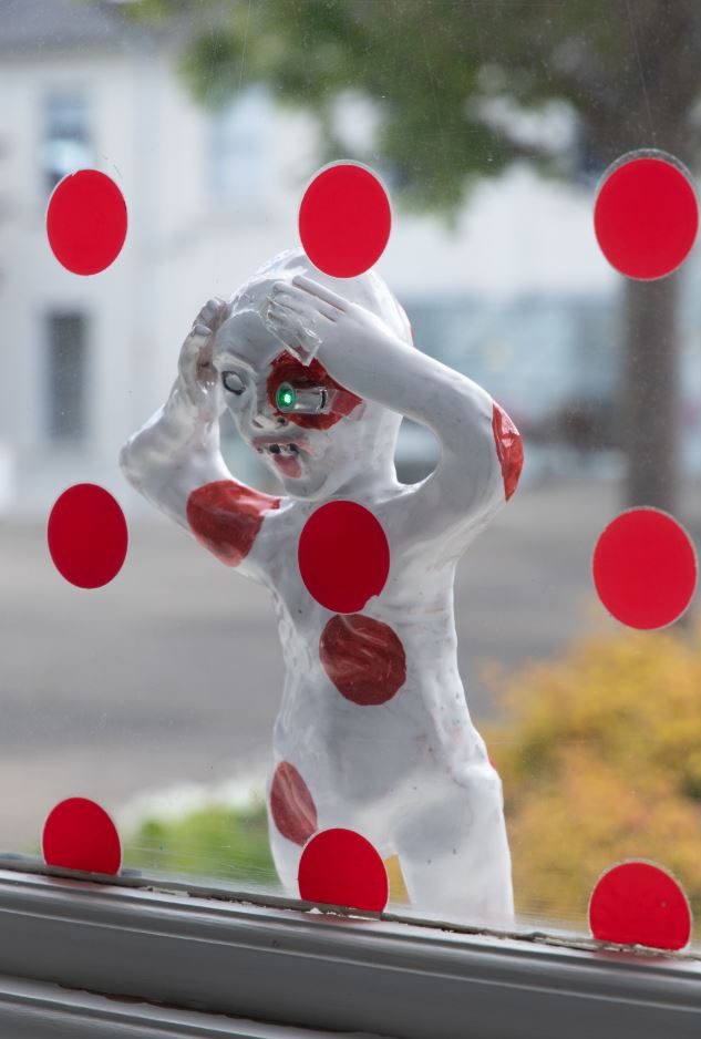 Image of a Sheela-na-gig wearing red polka dots looking through a window with red polka dot stickers on it