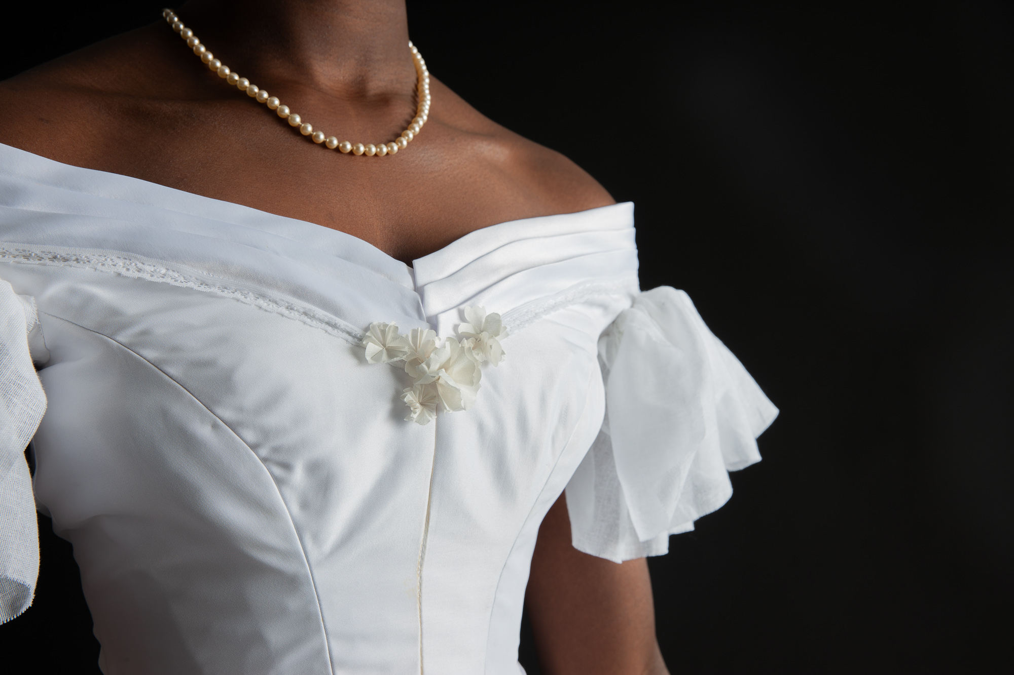 A close up of a white, off the shoulder bodice with short, ruffled sleeves, a pleated neckline, and small white flowers in the front being worn by a woman.