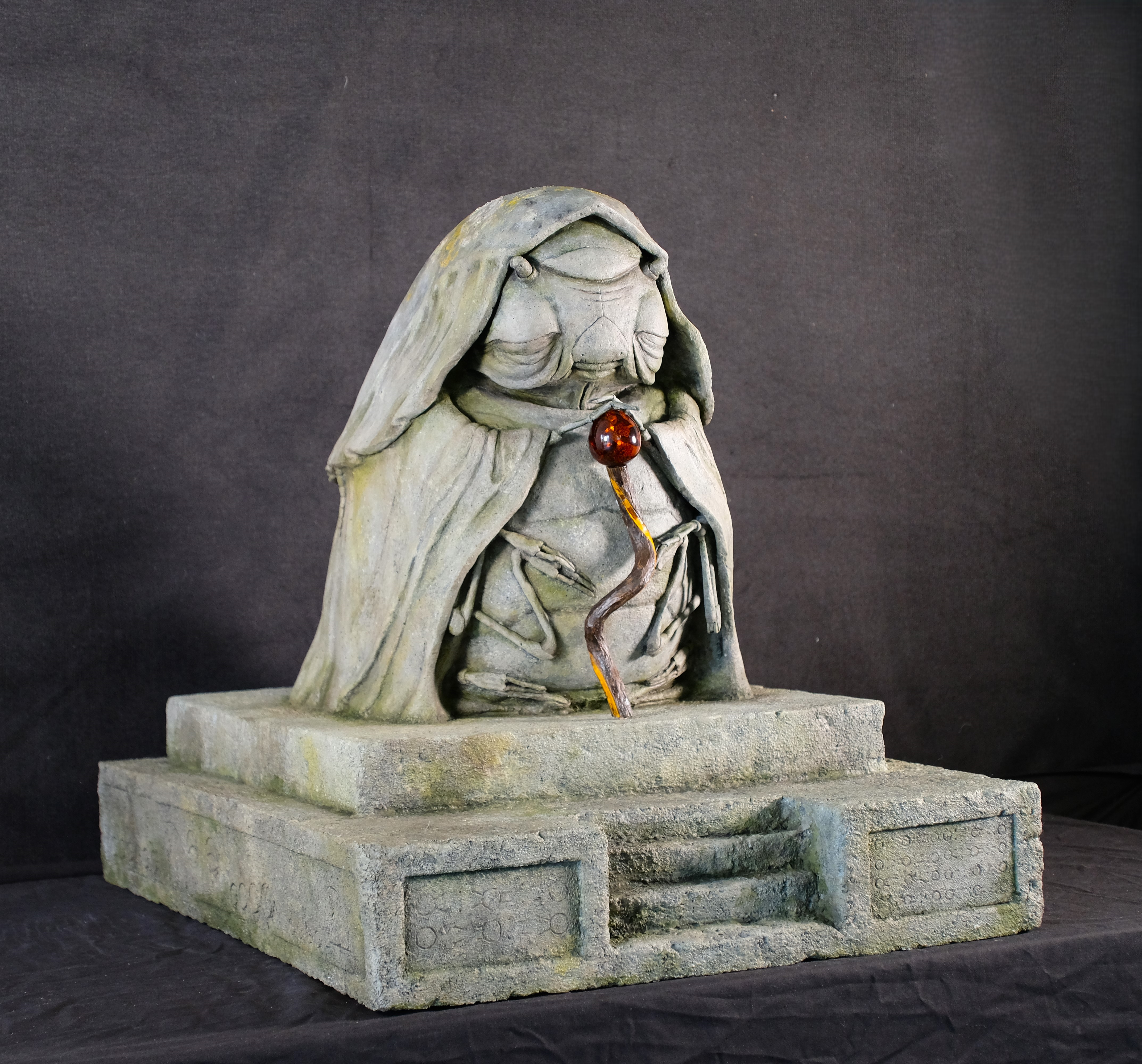 A photo of a stone-effect sculpture of an elderly lady/woodlouse hybrid, wearing a cloak and peacefully holding a wooden staff.