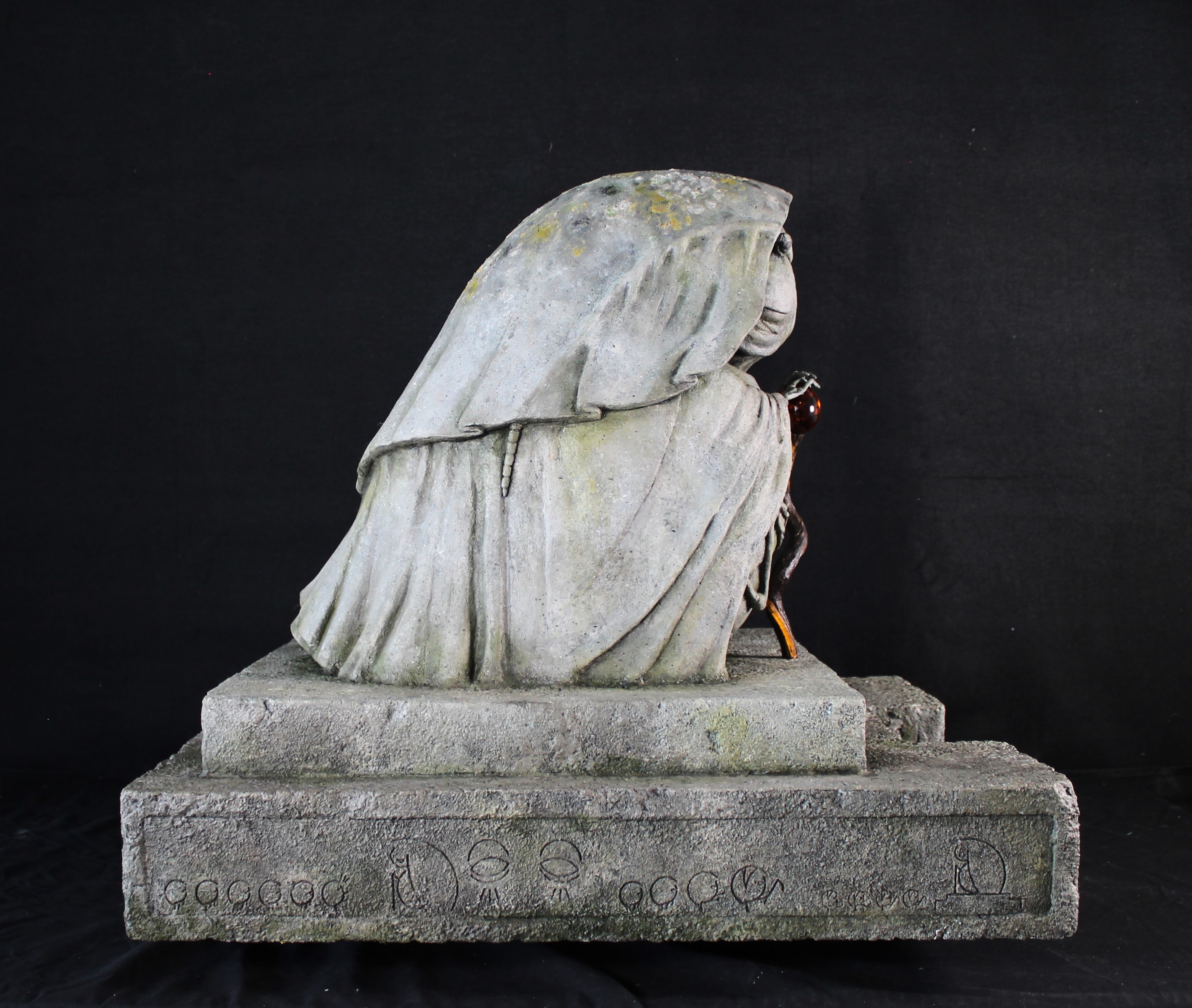 A photo of a stone-effect sculpture of an elderly lady/woodlouse hybrid, wearing a cloak and peacefully holding a wooden staff.