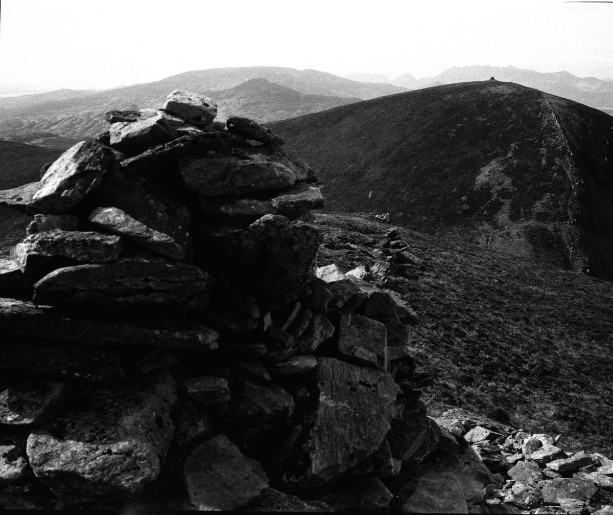 A black and white photograph of a mountainous landscape, featuring a cairn made of stacked stones in the foreground. Rolling hills and rugged peaks stretch into the background, with a steep path visible along the ridge of a prominent hill. The atmosphere is stark and dramatic, emphasizing the natural textures and contours of the terrain.