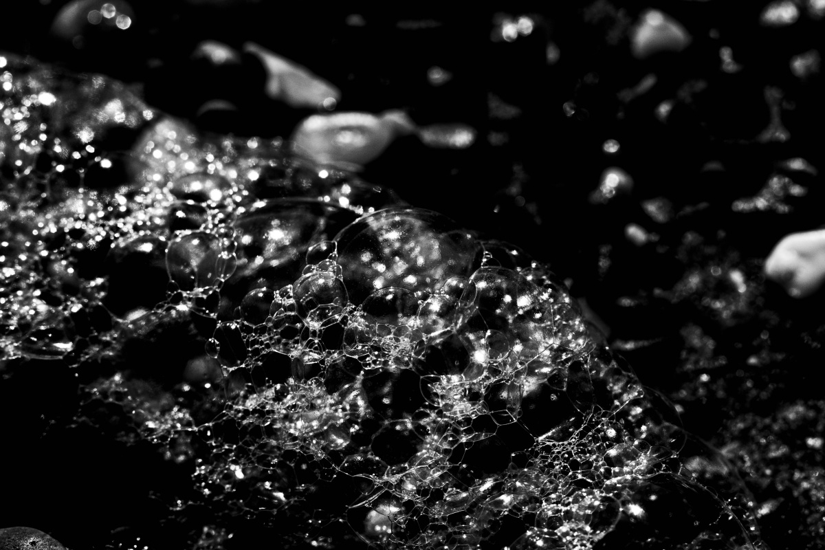 Black and white image taken on a digital camera depicting seafoam on  a beach. The bubbly outline of the foam starts on the left hand side of the photo and tapers off in the right bottom foreground. The grittiness of the sandy beach can also be seen in background and right side of the photograph.