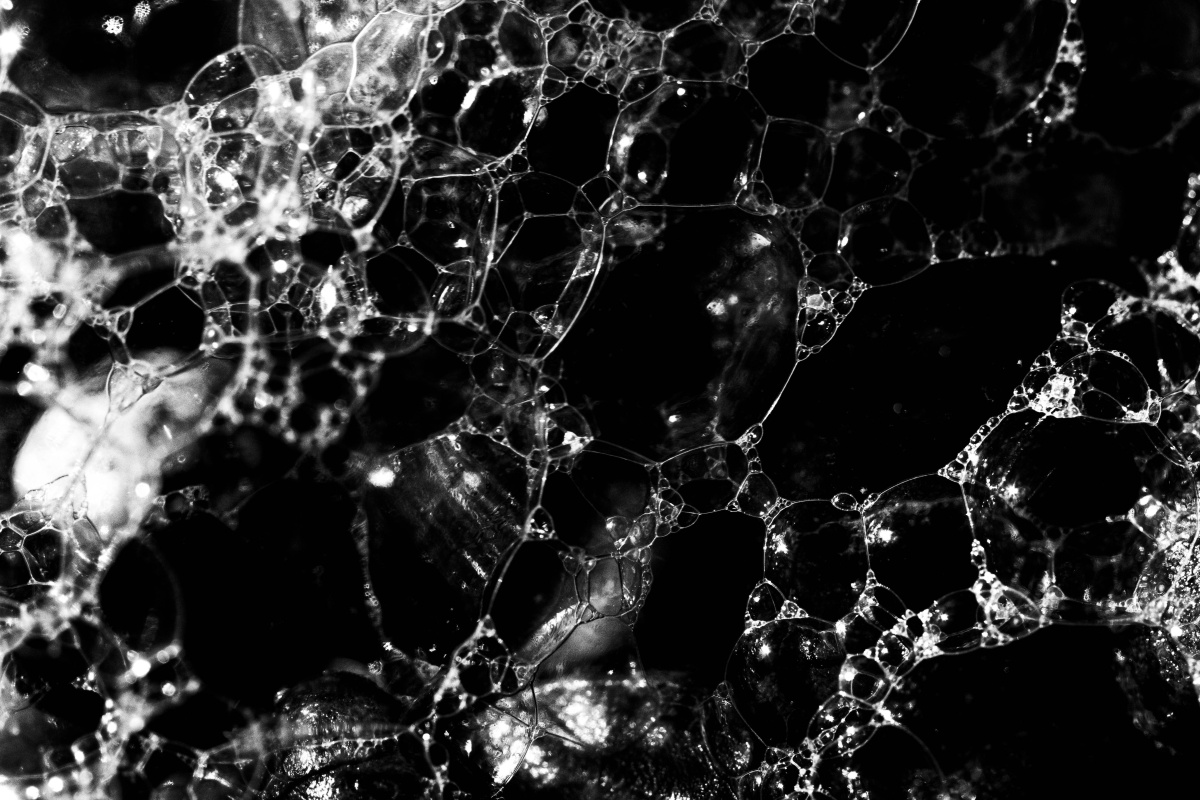 An even closer black and white macro shot of seafoam taken on a digital camera. In the top left corner there are a lot of white lines and grey spots on top of a black background that indicate a lot of bubbles clumped closely together. As you reach the centre and right side of the image the white lines become stretched further apart and there is not much grey, meaning the bubbles are bigger and aren't reflecting much light and you can see more of the black background