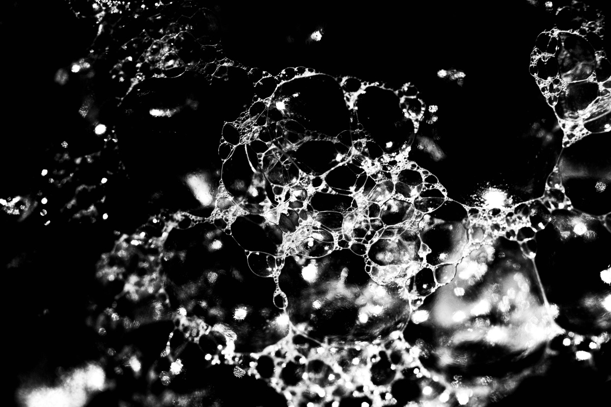 A black and white macro shot of seafoam taken on a digital camera. This image provides a closer look at the bubble-like formations of seafoam. It shows the white outlines of the bubbles enlarged further across the image from the left hand side to the right. In the middle of the bubbles we can see the blackness of the background and some grey-white smudges where the light reflected off the foam, showing off the texture of the bubbles. 