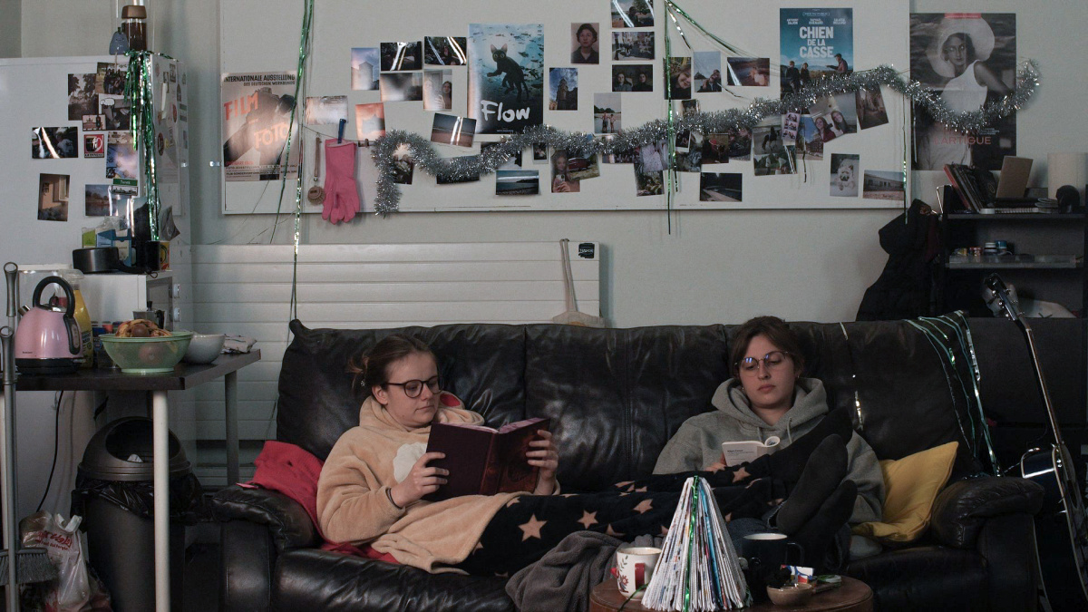 The photograph shows two young women sitting on a black leather sofa. They are sitting in the middle of a messy living room. The young woman on the left is reading a book. She is wearing reindeer pyjamas and glasses. The woman on the right, wearing a grey hoodie and glasses, is also reading a book. They are not looking at each other, but sharing a moment together.