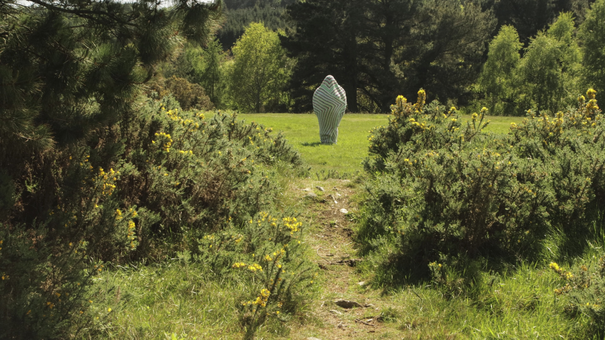 Logan Valentine in a Crocheted Cocoon, in a field in the distance, framed by bushes and trees