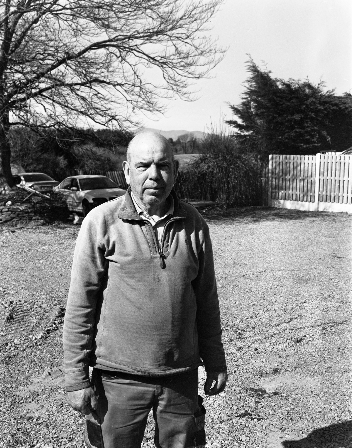 A black and white photograph of a person standing outdoors on a gravel surface. The individual is wearing a zip-up sweater and work trousers with reinforced knees. In the background, there are leafless trees, a wooden fence, and a couple of parked cars partially visible among bushes. The scene is brightly lit, suggesting it is daytime, and there are hills visible in the distance. A cigarette sits hidden in his left hand.
