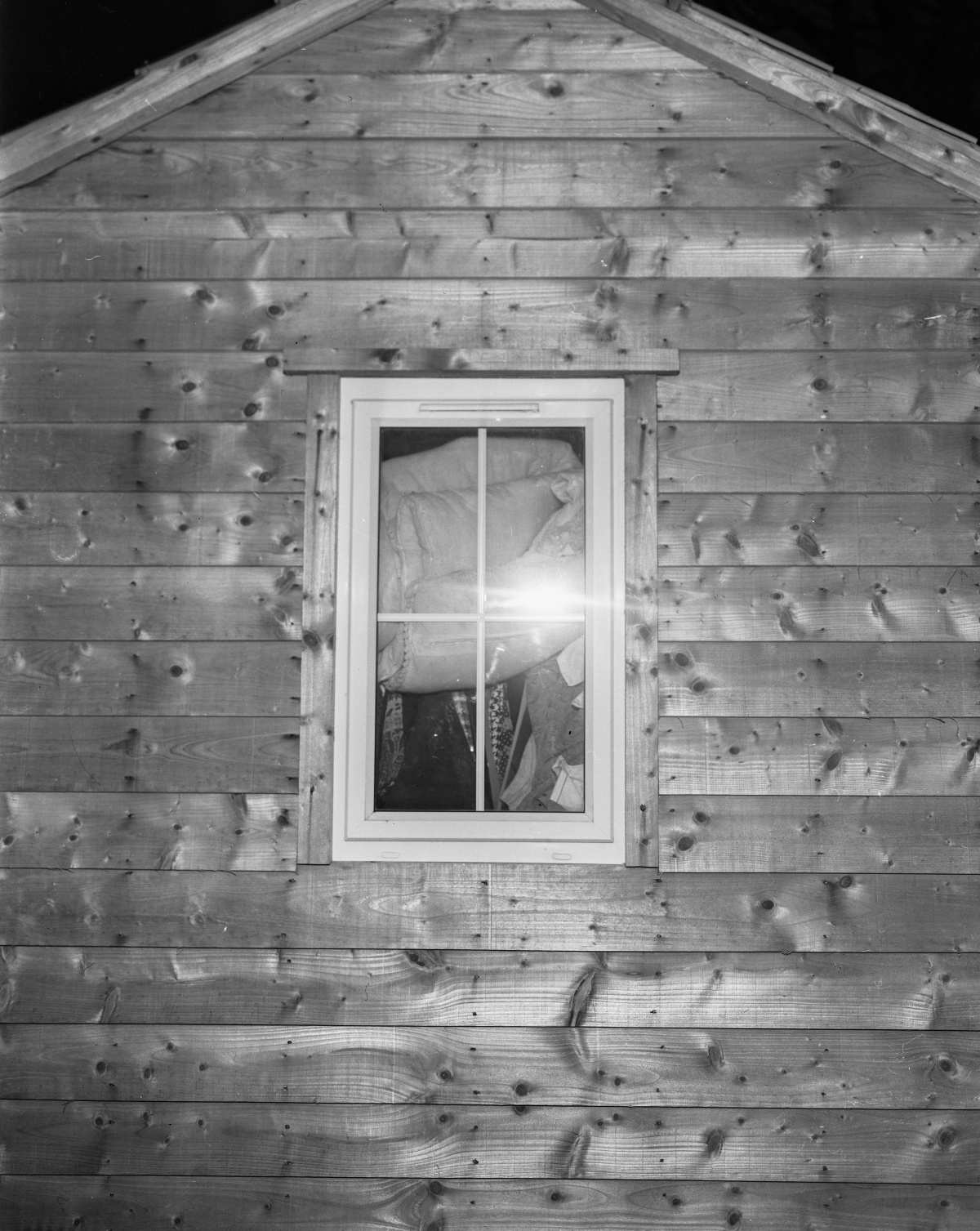 A black and white photograph of the exterior wall of a wooden shed, viewed straight on. In the center is a single window with a white frame, through which various bulky items, such as bags or blankets, are piled up and pressed against the glass. The camera flash creates a bright glare on the window, partially obscuring the view inside. The wood grain and knots in the shed’s planks are clearly visible, giving the image a textured appearance.