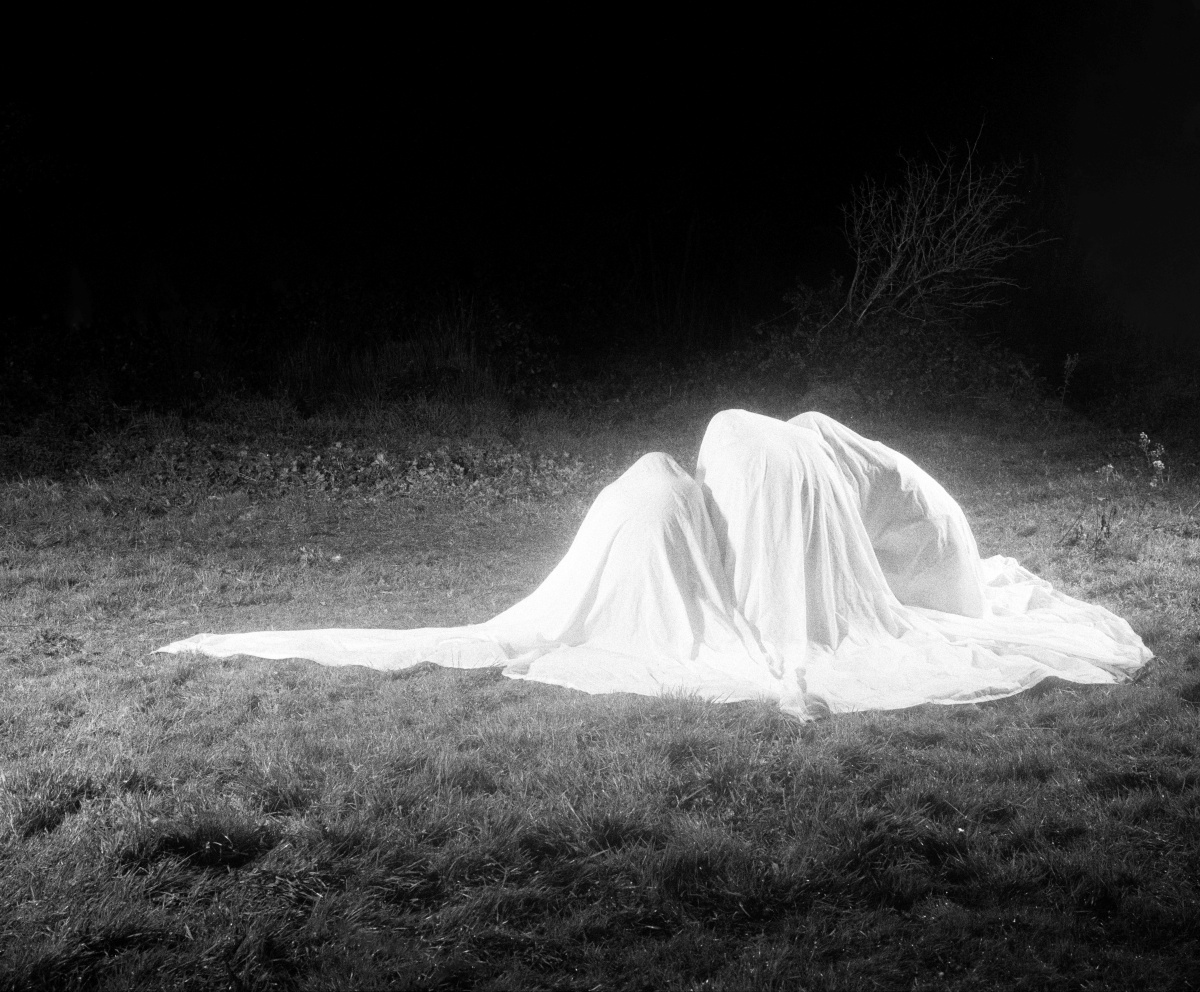 A black and white photograph depicting three human-like forms covered in a large white sheet,  kneeling on a grassy field at night. The scene is dramatically lit, casting a glow on the sheet and surrounding grass while the background fades into darkness. Sparse vegetation and a bare shrub are visible at the edge of the illuminated area, adding to the eerie and surreal atmosphere.