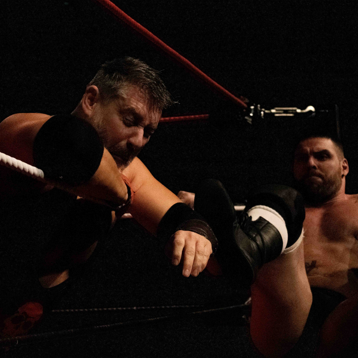 One man is lying against the ropes, bracing for the impact from the kick to the head from the second man