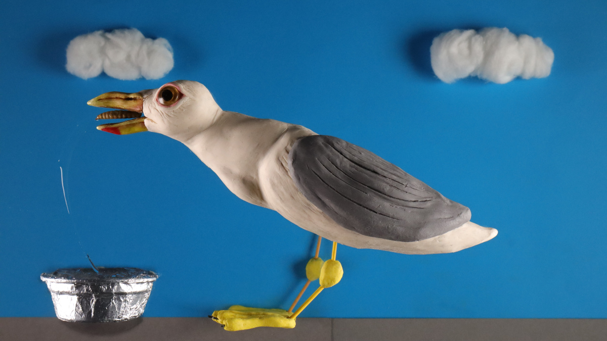 A still of a claymation scene on a multiplane. A plasticine seagull is mid chomp on a dried mealworm from a tinfoil dog bowl, with a trail of hair gel representing a water splash. 