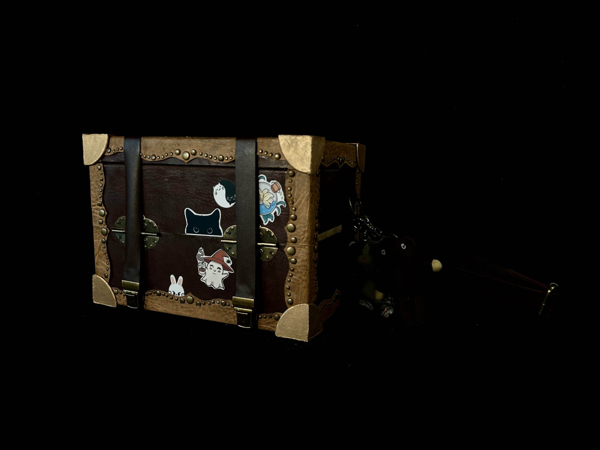A Closed leather-bound box, with a shoulder strap. The straps are closed. There are stickers of ghosts, cats, and a bunny on the front, and two keychains of a cat and a monkey. on the side 