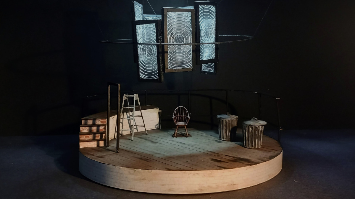 Miniature scale model of a circular stage, with large fresnel lens arranged in a circle hanging above the stage. On the stage there is a metal stepladder, a wooden chair, and two metal bins. Blue light shines through the fresnel lenses.