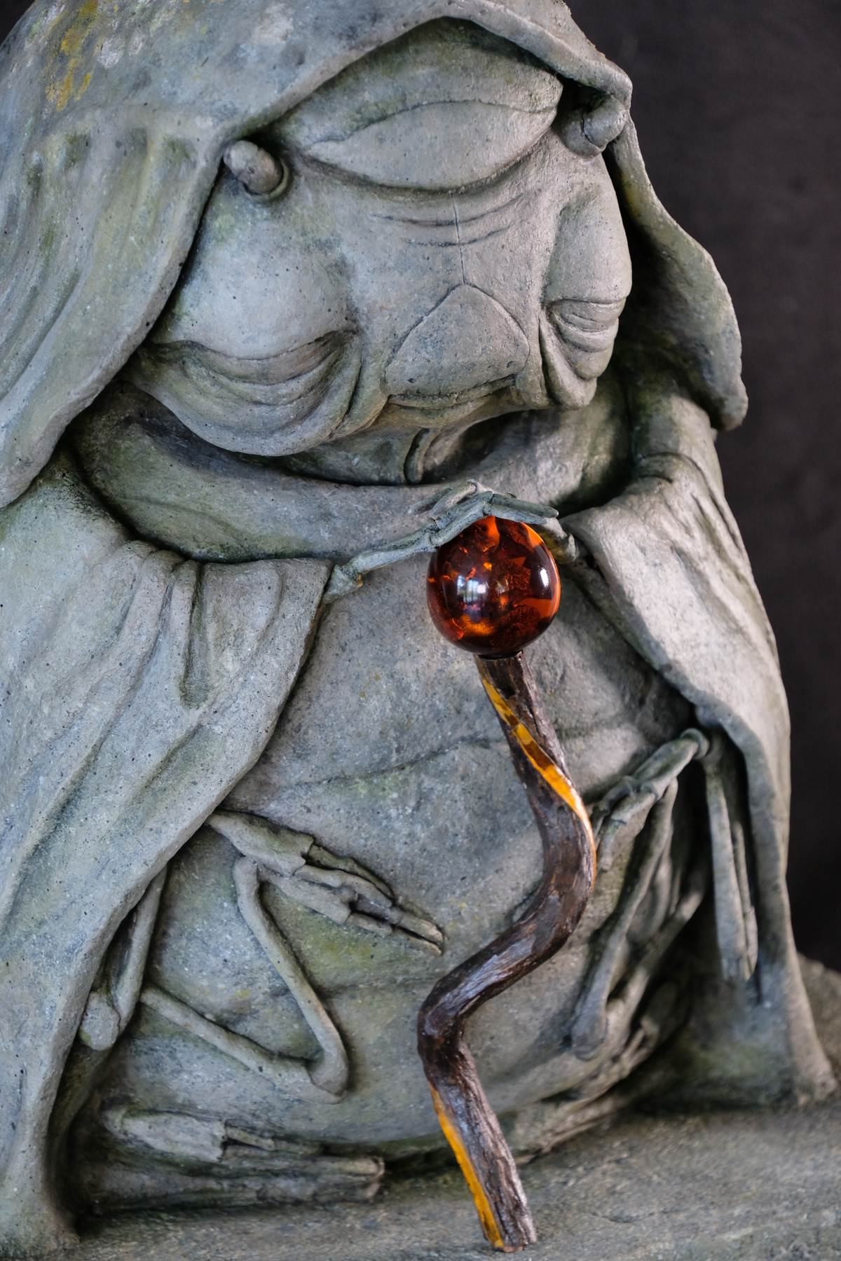 A photo of a stone-effect sculpture of an elderly lady/woodlouse hybrid, wearing a cloak and peacefully holding a wooden staff.
