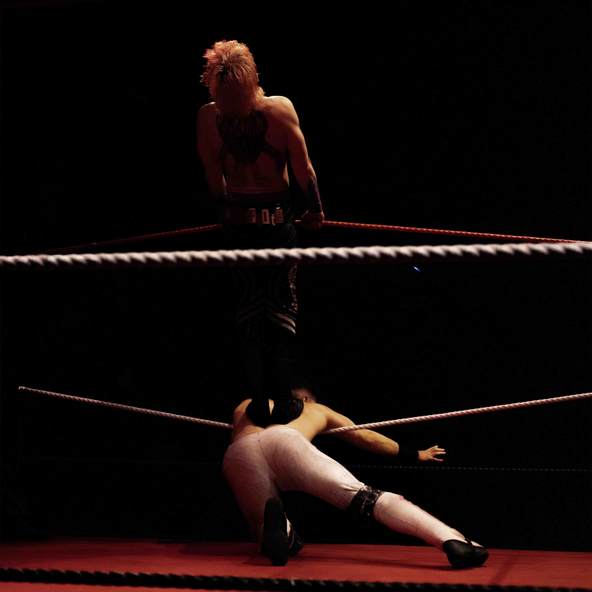 A man standing on the back of another man as he pushes him down between th eropes of a wrestling ring