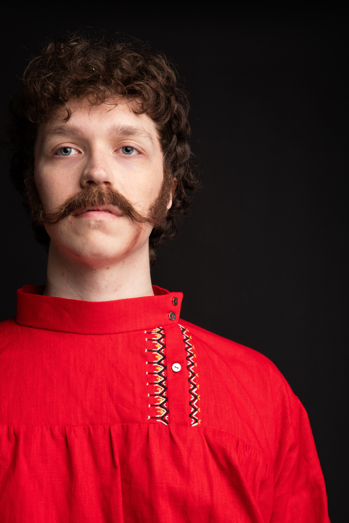 A man with sideburns and moustache in a red kosovorotka peasant shirt with black, white, and yellow embroidery standing facing the viewer.