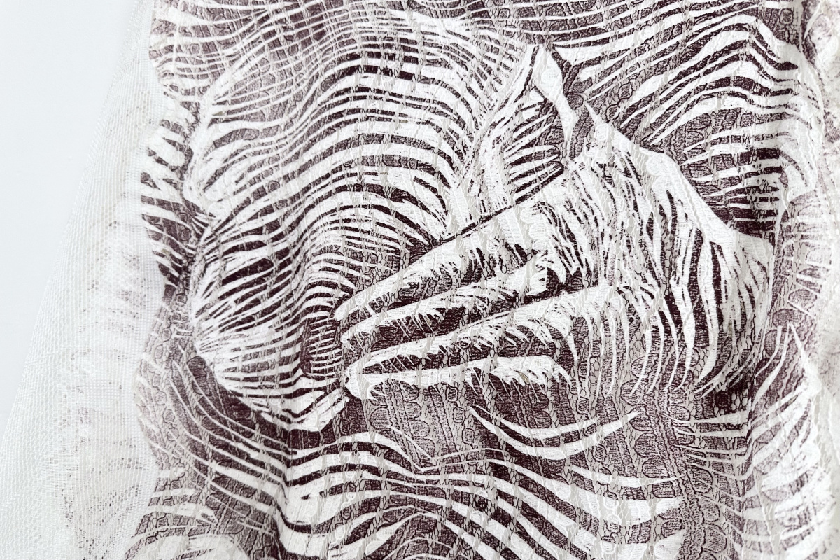 A closeup of a print on a lace dress. A hand is depicted covering a woman's torso, with the lace underneath texturing the print.