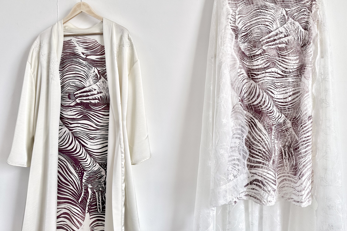 A closeup photo of two pieces of clothing hanging from the wall. Both have the same print of a woman’s body done with striped line. She is covering herself with both hands. The piece on the left is a yellowish robe, the one on the right a lacy white dress with more lace hanging from it.