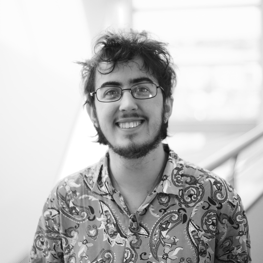 A black and white photograph of the artist. Theo is white, has short hair, rectangular glasses, a beard and patterned short-sleeved button up shirt. They're smiling