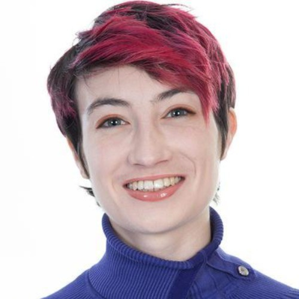 Mary Elizabeth Mc Grath Headshot. White woman with pink pixie cut and blue turtle neck.