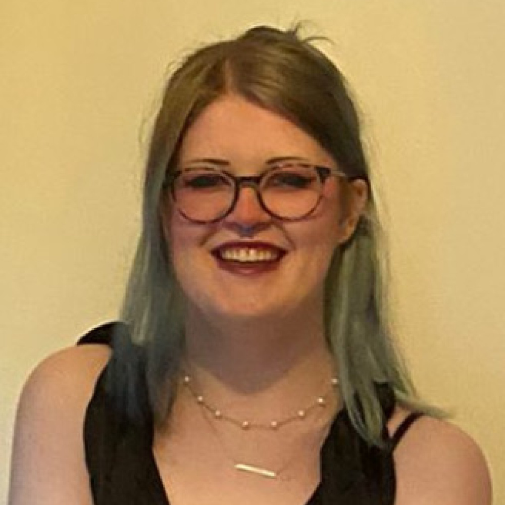 Smiling woman with brown and light green ends hair, wearing round glasses, two necklaces with the wide straps of a black dress visible on the shoulders