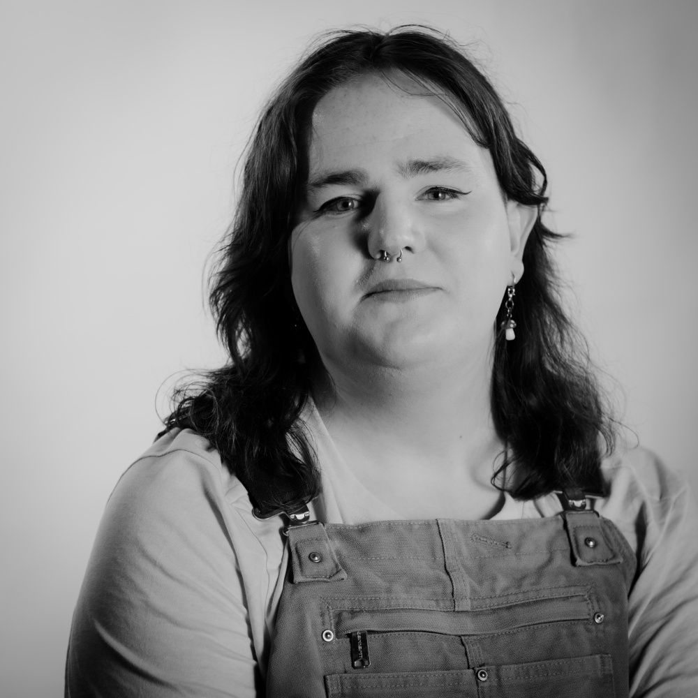 Casey is seen smilling, wearing dungarees and a light t-shirt, against a white background.