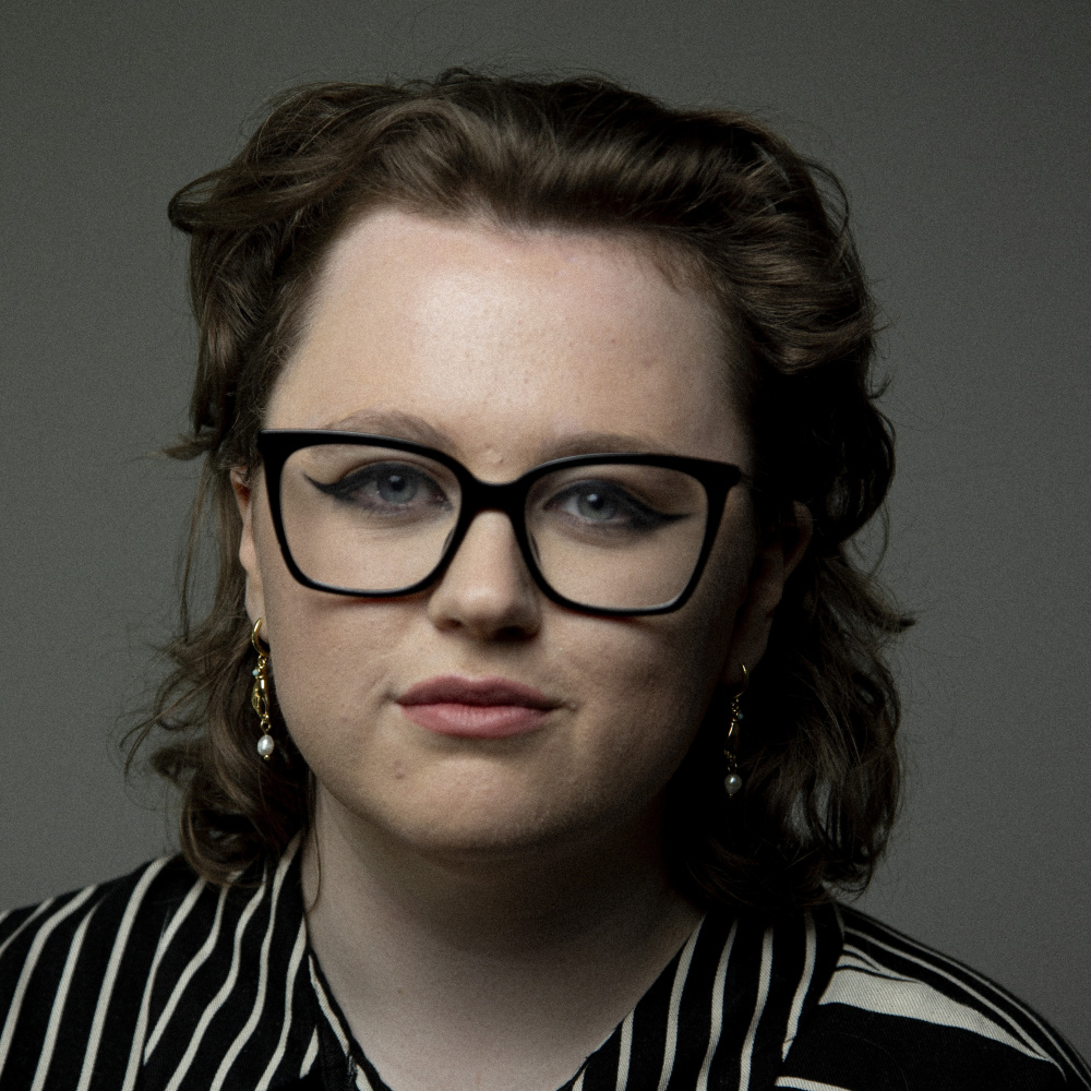 Profile picture of Ciara. Wearing square black frame glasses, black and cream stripe shirt, and gold hand with pearl earrings. Ciara has a brown curly shoulder length mullet. Round features, sharp makeup such as eyeliner and a slight cheeky grin.