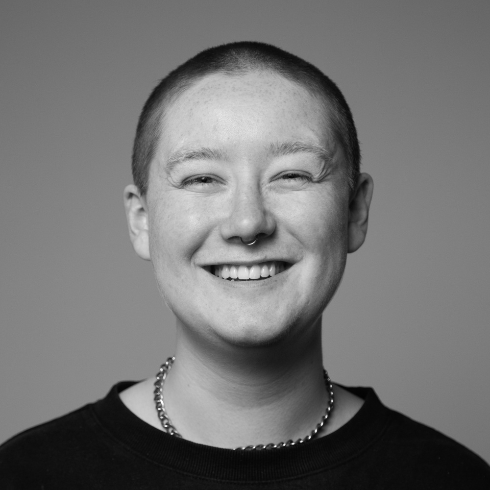 woman in their early 20's shaved head, freckled, wide smile, wearing a small chain and a black t-shirt
