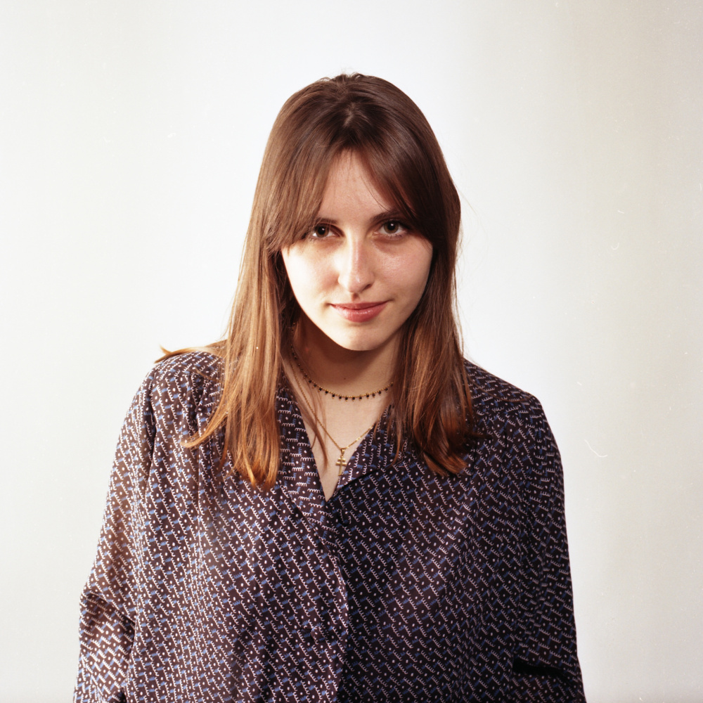 Analogue photography of a young woman looking directly at the lens of the camera. She has slightly long hair, brown eyes and is wearing a blouse. She is smiling.