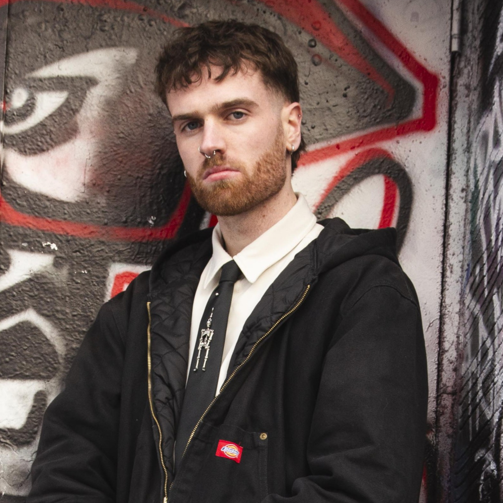 Photo of a man in a shirt, tie and black jacket. The tie has a skeleton attached to it. The man is standing in front of a black and red graffitied wall