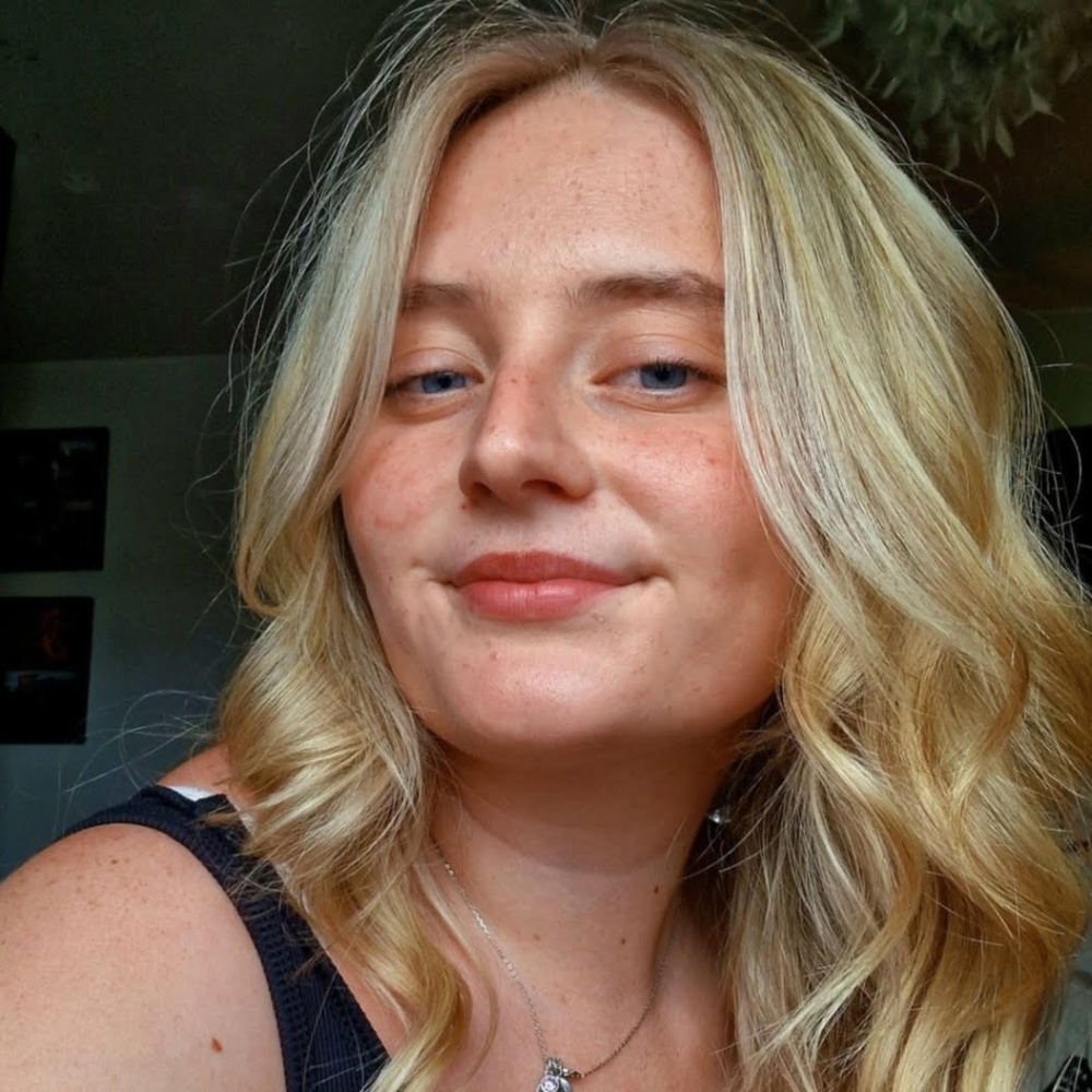Close-up portrait-style selfie of a young woman with long, wavy styled blonde hair and light freckles with blue eyes with a relaxed, slight smile expression. She is wearing a sleeveless black top with a small necklace with a round pendant.