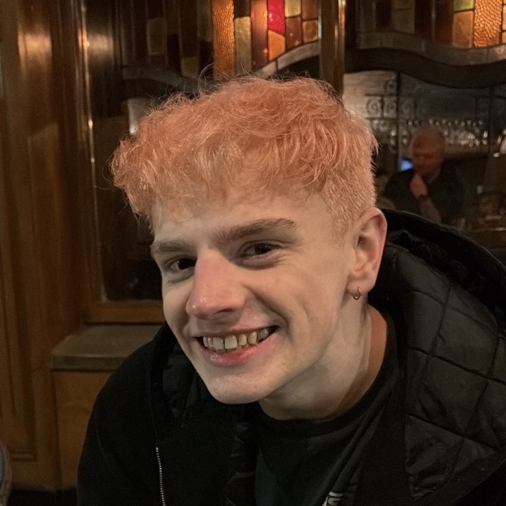 Padraig Gilmore smiling, he has light pink hair and hazel eyes.