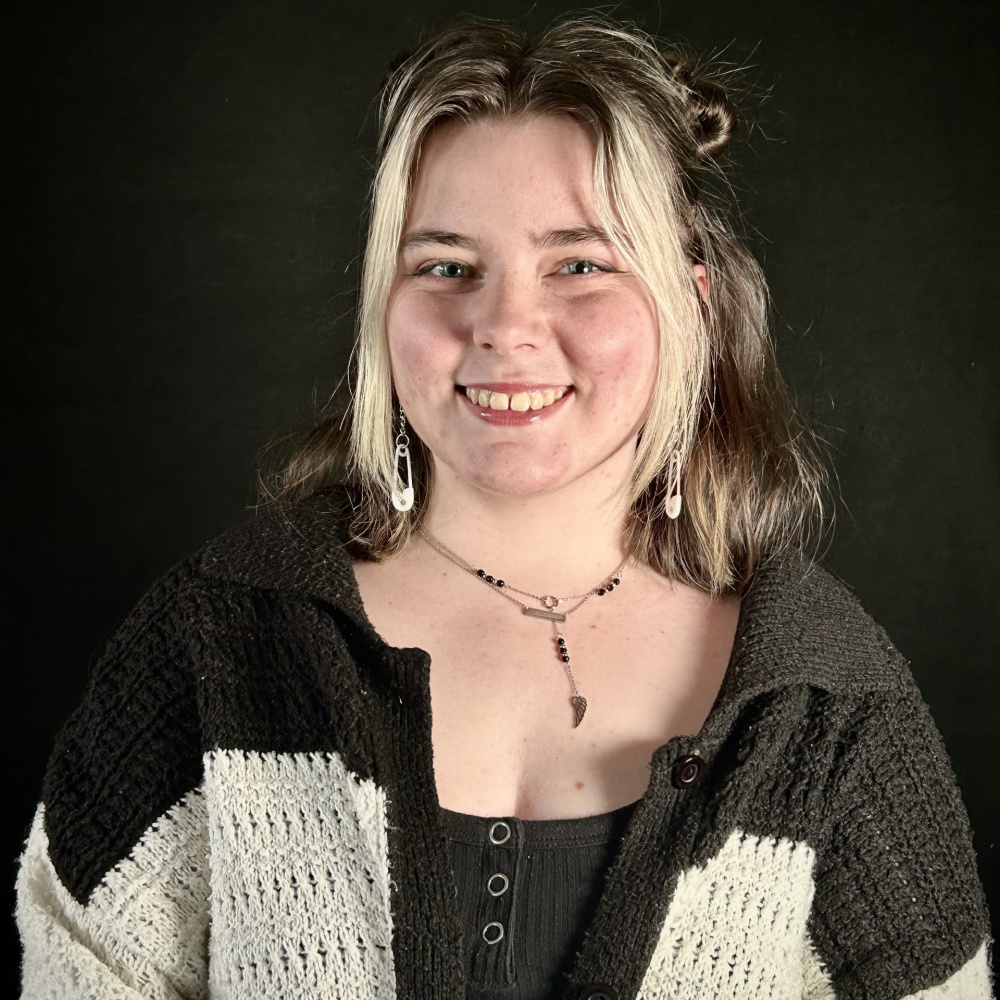 White woman standing in front of a black background. She has brown hair with blonde money pieces in the front. Her hair is half up-half down with buns. She is smiling, and wearing a black tank top and a black and white striped cardigan